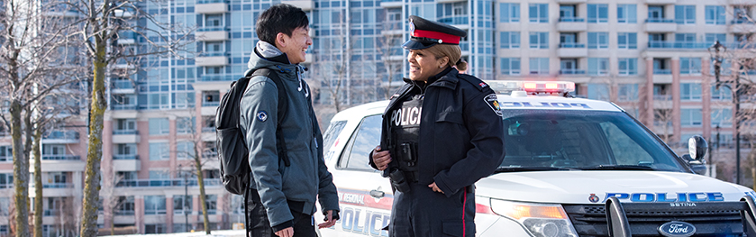 A woman in a police uniform smiles and speaks to another man in front of a police cruiser
