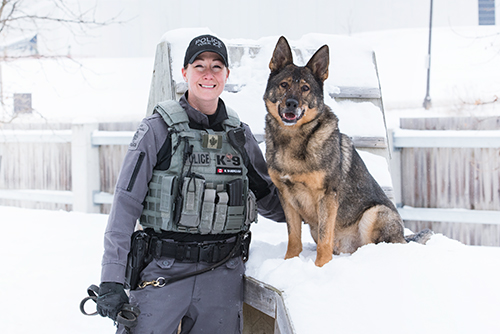 A woman in a police uniform smiles next to a large dog, in a snowy yard.