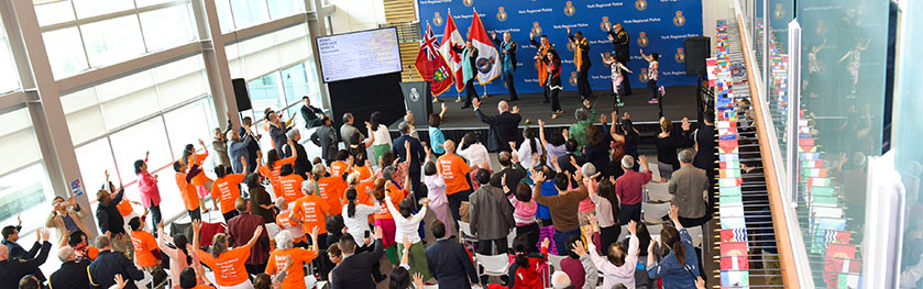 A group of five adults and two kids dance on stage in front of a large crowd at police headquarters
