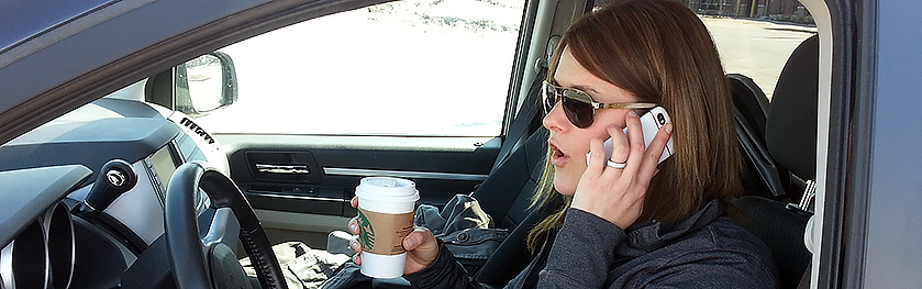 A woman talks on her phone and holds a coffee while driving