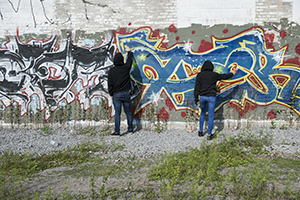 A pair of people in black hoodies spray paint a wall