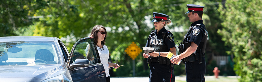 A woman leans against a car while speaking with two people in police uniforms