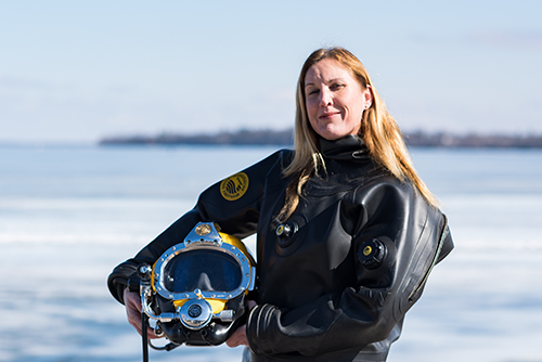 A blonde woman stands in front of a frozen lake in a black rubber suit.