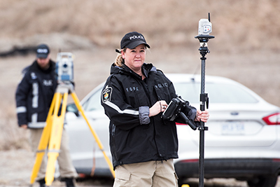 A woman in a police jacket and khaki pants writes on a tablet in front of a car