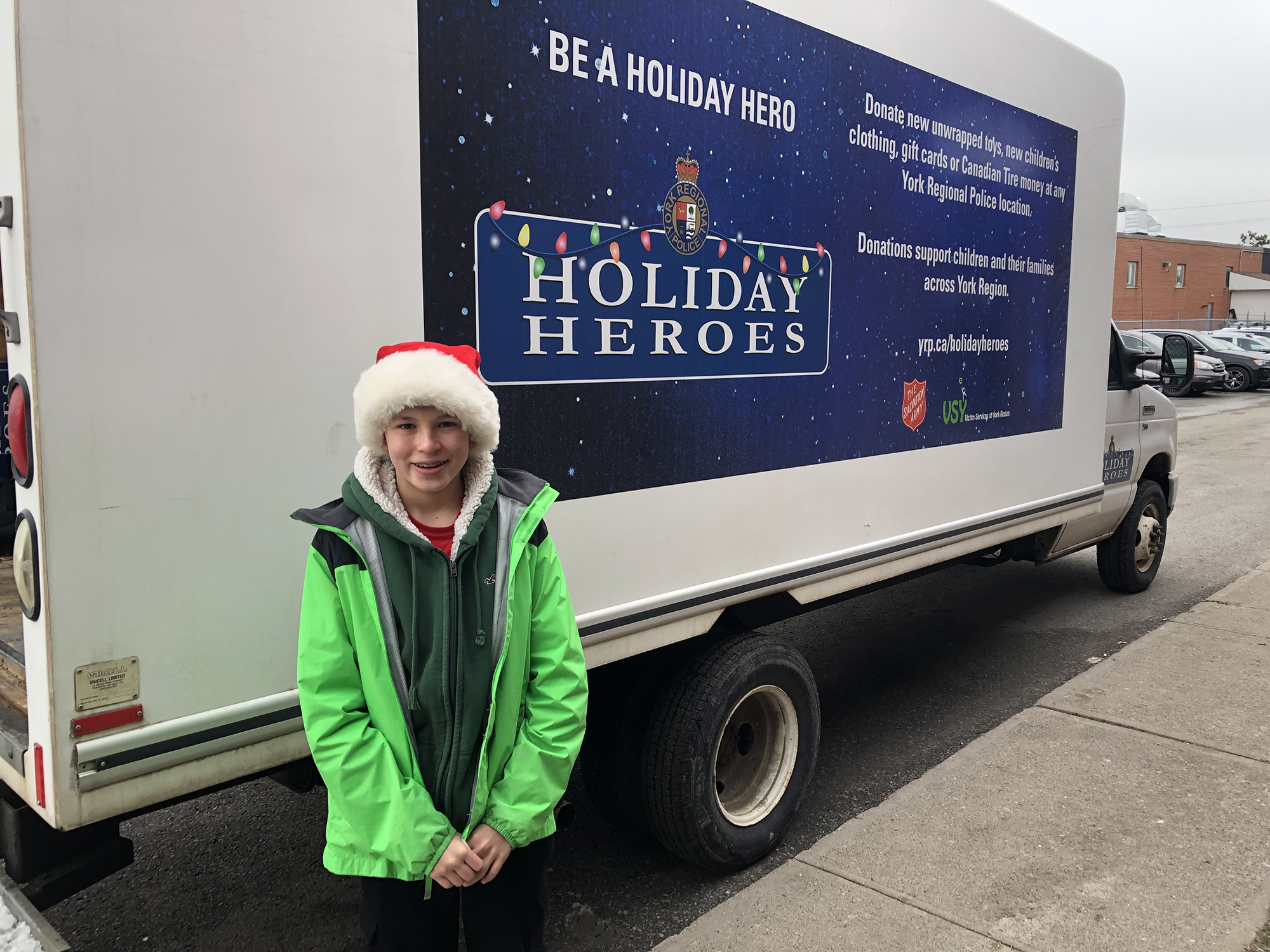 A male student in a Santa hat stands in front of the Holiday Heroes truck