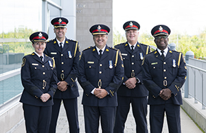 A group of four men and one woman in formal police uniforms, smiling on a sunny day
