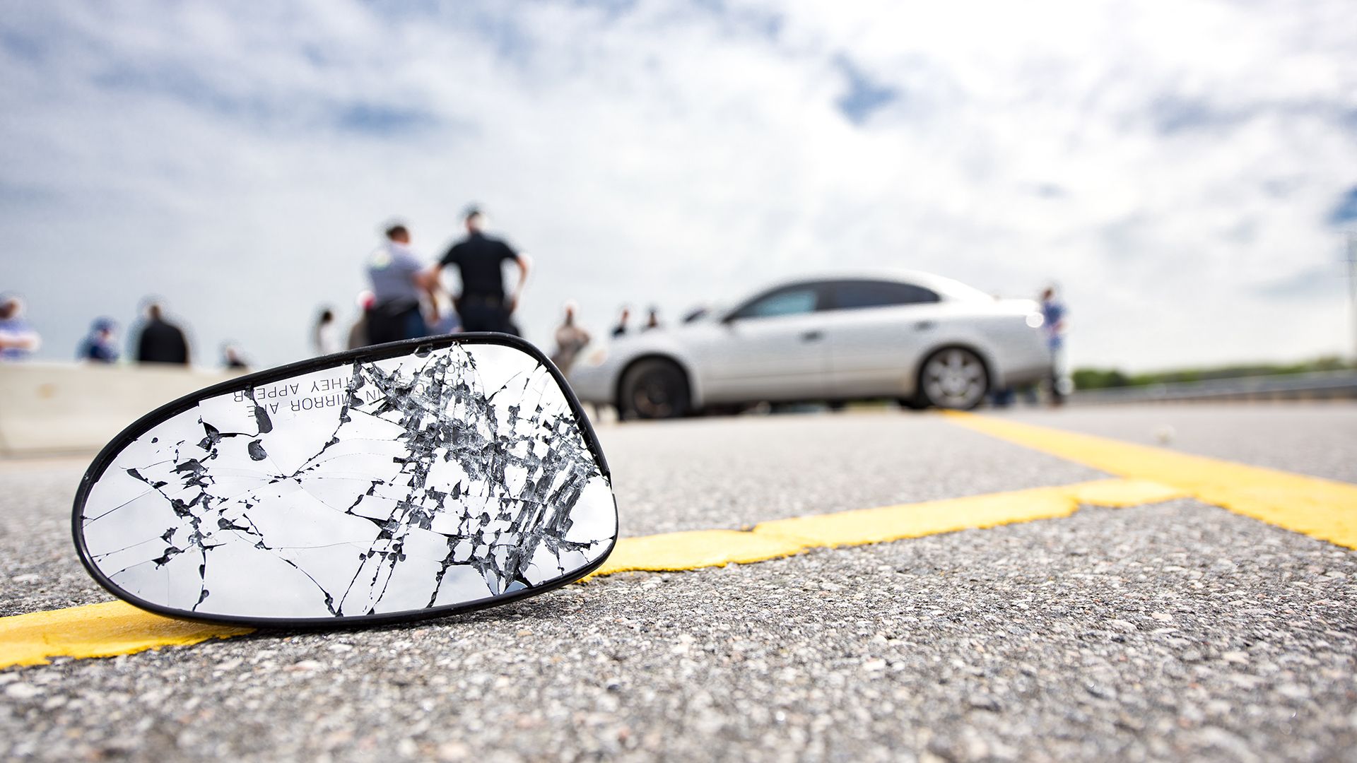 A broken car mirror is seen on the pavement in front of a motor vehicle collision
