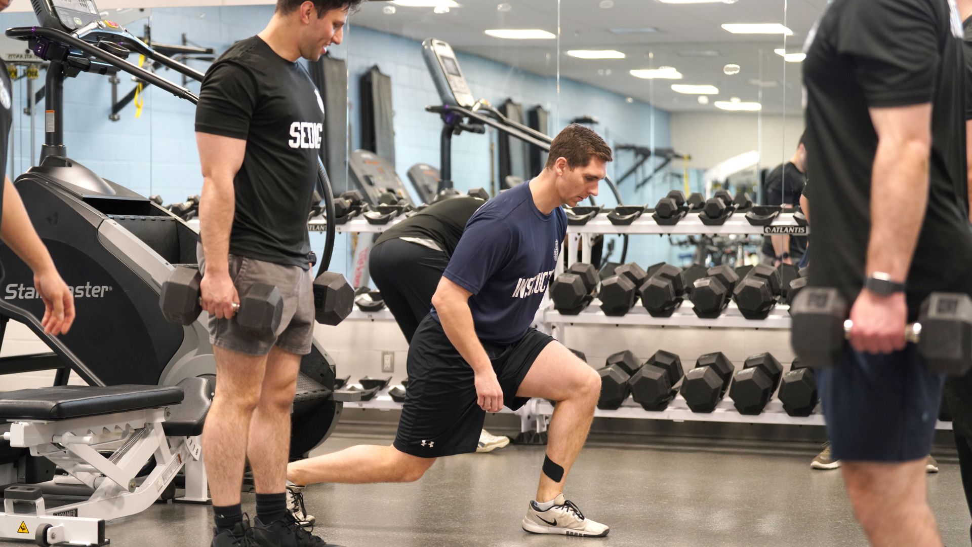 A man observes another man performing a lunge in a gym. 