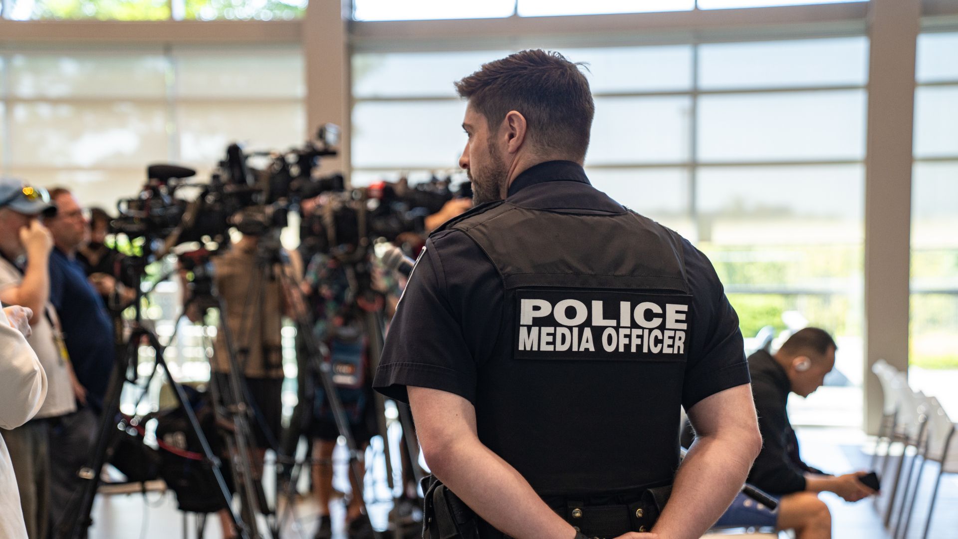 An officer wearing a vest that says police media officer walks in front of cameras at a press conference