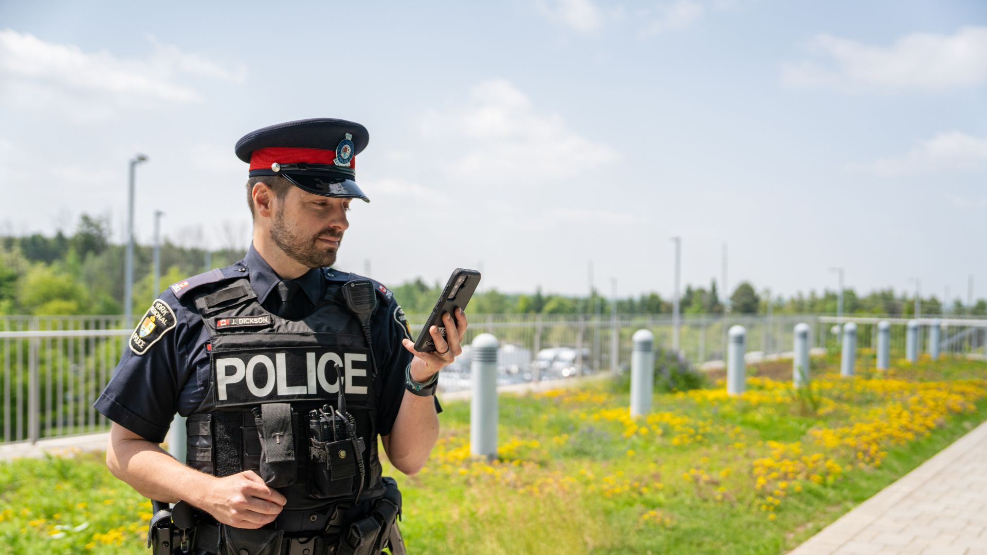 A police officer in uniform stands outdoor in a park looking at a cellphone.