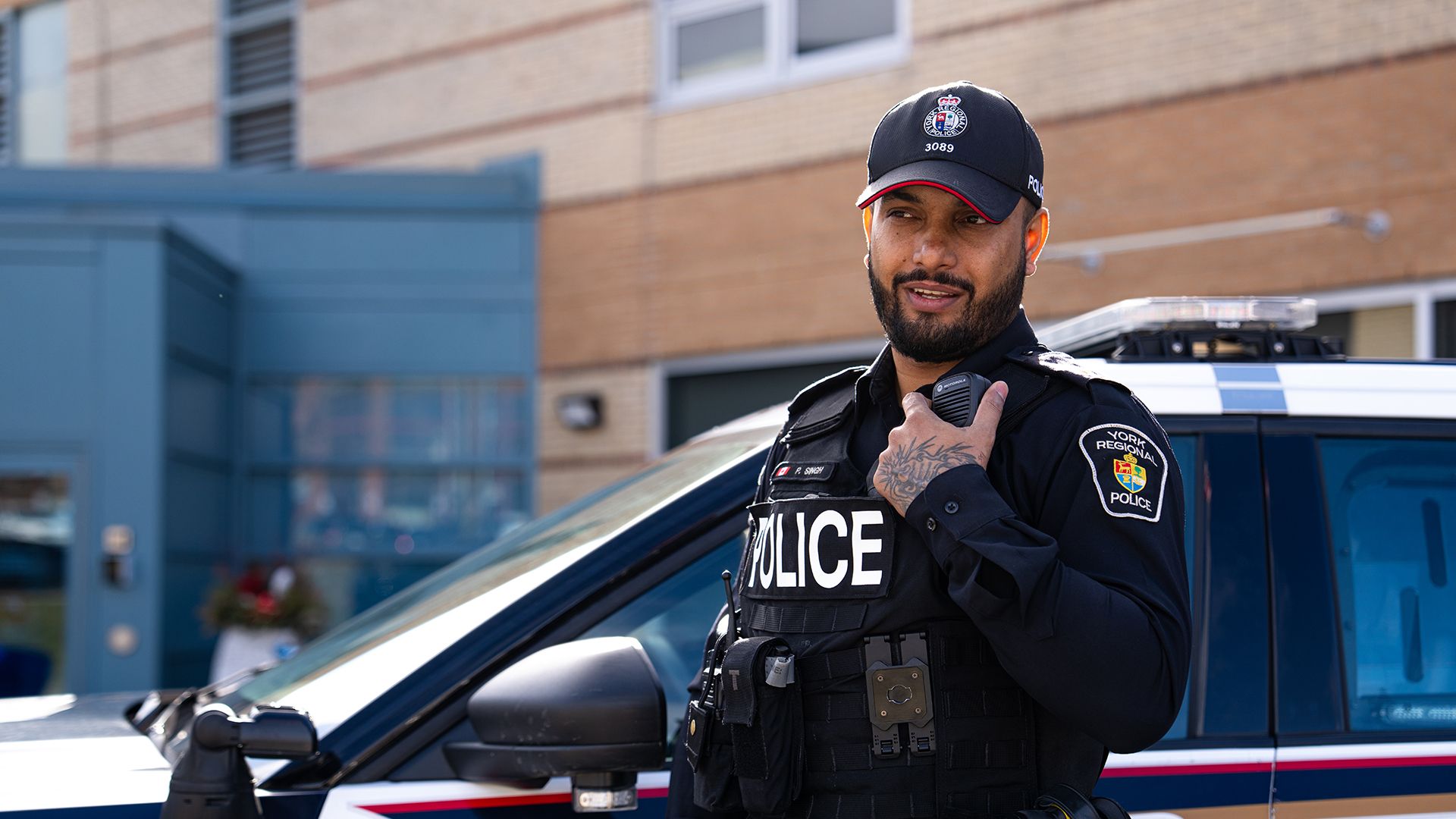 Police officer stands next to a police cruiser speaking into a handheld radio.
