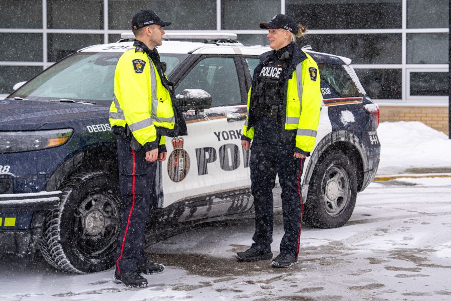 Two police officers in reflective jackets stand in front of a cruiser in snowy weather.