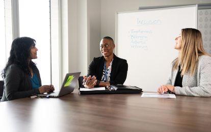 Three people sit in a board room in front of a white board that reads INCLUSION
