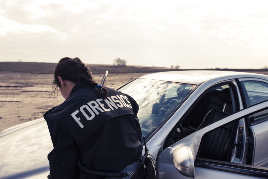 A person in a jacket that reads FORENSICS examining the hood of a car.