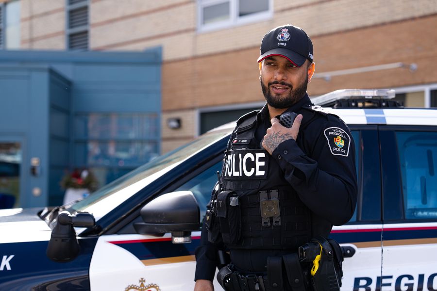 Police officer stands next to a police cruiser speaking into a handheld radio.