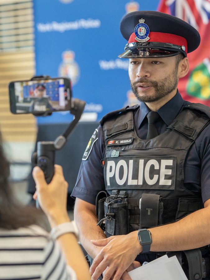 A man in a police uniform stands in front of a woman recording on a phone