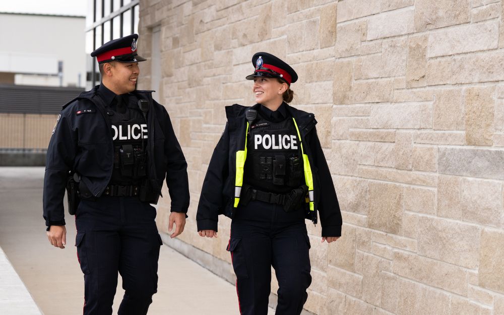 Two police officers walk and chat along a brick building.